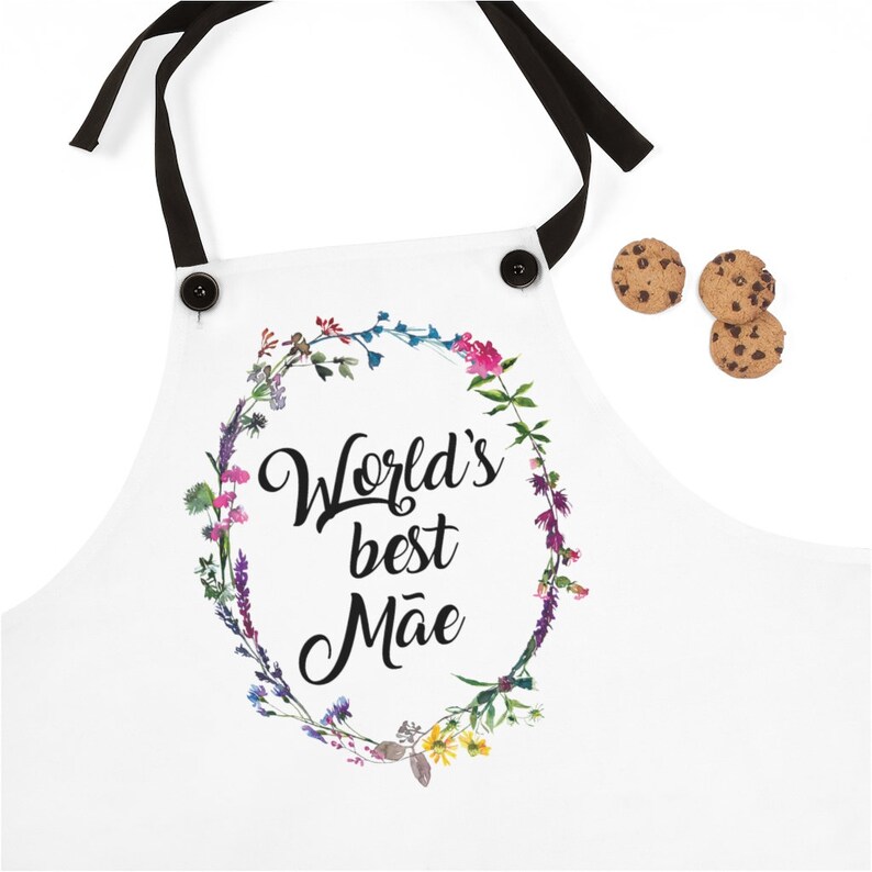 May include: White apron with black straps and buttons. The apron has a floral wreath design with the text "World's best M&atilde;e" in black.