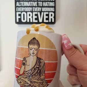 K&ouml;nnte beinhalten: Eine wei&szlig;e Keramiktasse mit einer Grafik eines meditierenden Buddhas vor einem Sonnenuntergangs-Hintergrund. Die Tasse hat den Text "Let that shit go" auf dem Boden aufgedruckt. Die Tasse ist mit einem Kaffeegetr&auml;nk gef&uuml;llt, das mit Schlagsahne und Karamell betr&auml;ufelt ist.