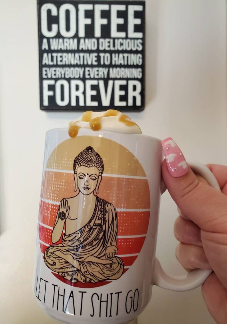 K&ouml;nnte beinhalten: Eine wei&szlig;e Keramiktasse mit einer Grafik eines meditierenden Buddhas vor einem Sonnenuntergangs-Hintergrund. Die Tasse hat den Text "Let that shit go" auf dem Boden aufgedruckt. Die Tasse ist mit einem Kaffeegetr&auml;nk gef&uuml;llt, das mit Schlagsahne und Karamell betr&auml;ufelt ist.