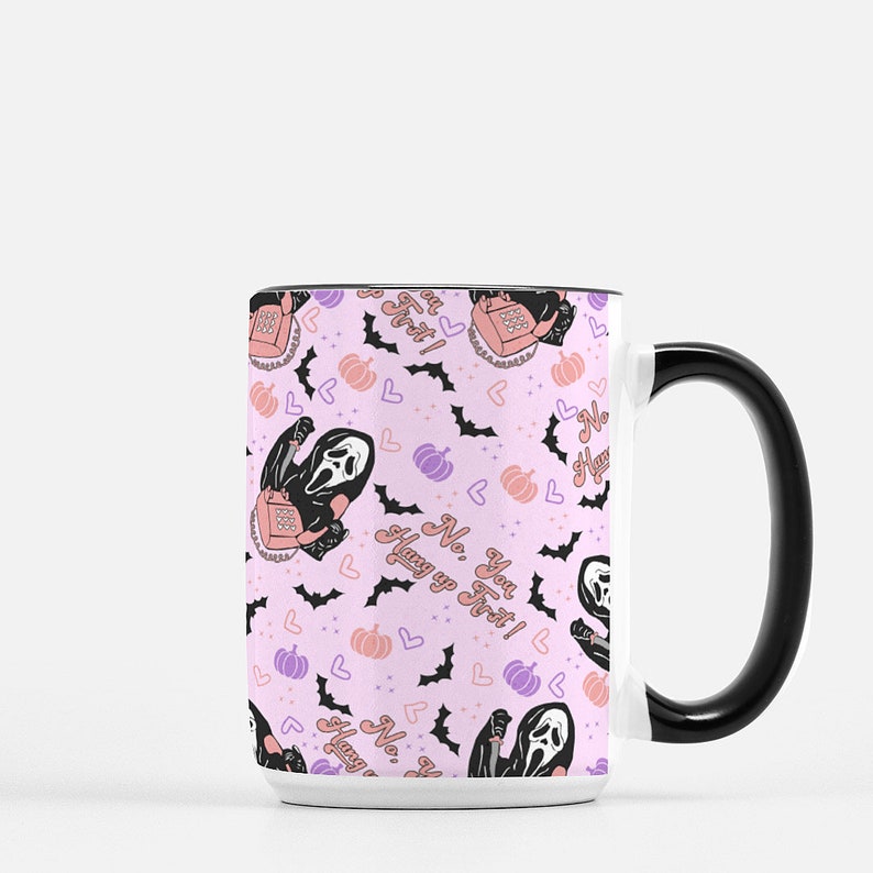 May include: White ceramic mug with a black handle. The mug features a pink background with a repeating pattern of a ghost holding a phone, bats, hearts, and pumpkins. The text "No, you hang up first!" is repeated throughout the pattern.