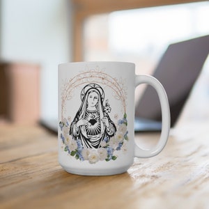 May include: White ceramic mug with a black and white image of the Virgin Mary with a heart and flowers. The mug has a gold floral design around the image.