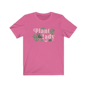 May include: Pink short-sleeved t-shirt with the text "Plant Lady" in a retro font. The text is surrounded by a variety of green plants, including cacti and succulents.