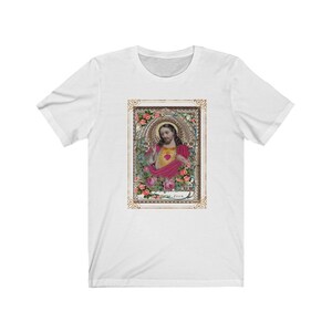 May include: White t-shirt with a vintage image of Jesus with a heart in his hand. The image is surrounded by a gold frame with floral details. The text "Jesus" is at the bottom of the image.