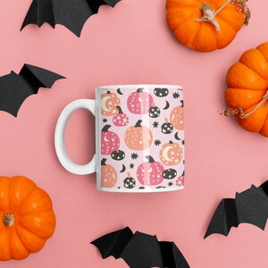 May include: A white ceramic mug with a pink and orange pumpkin pattern. The mug is surrounded by black paper bats and orange pumpkins on a pink background.