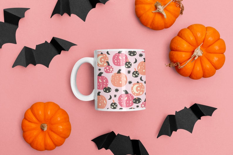 May include: A white ceramic mug with a pink and orange pumpkin pattern. The mug is surrounded by black paper bats and orange pumpkins on a pink background.