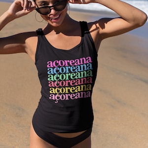 Op de afbeelding: Een zwart tanktop met het woord "acoreana" zes keer herhaald in verschillende kleuren: roze, blauw, groen, geel, oranje en roze. Het tanktop wordt gedragen door een persoon op een strand.