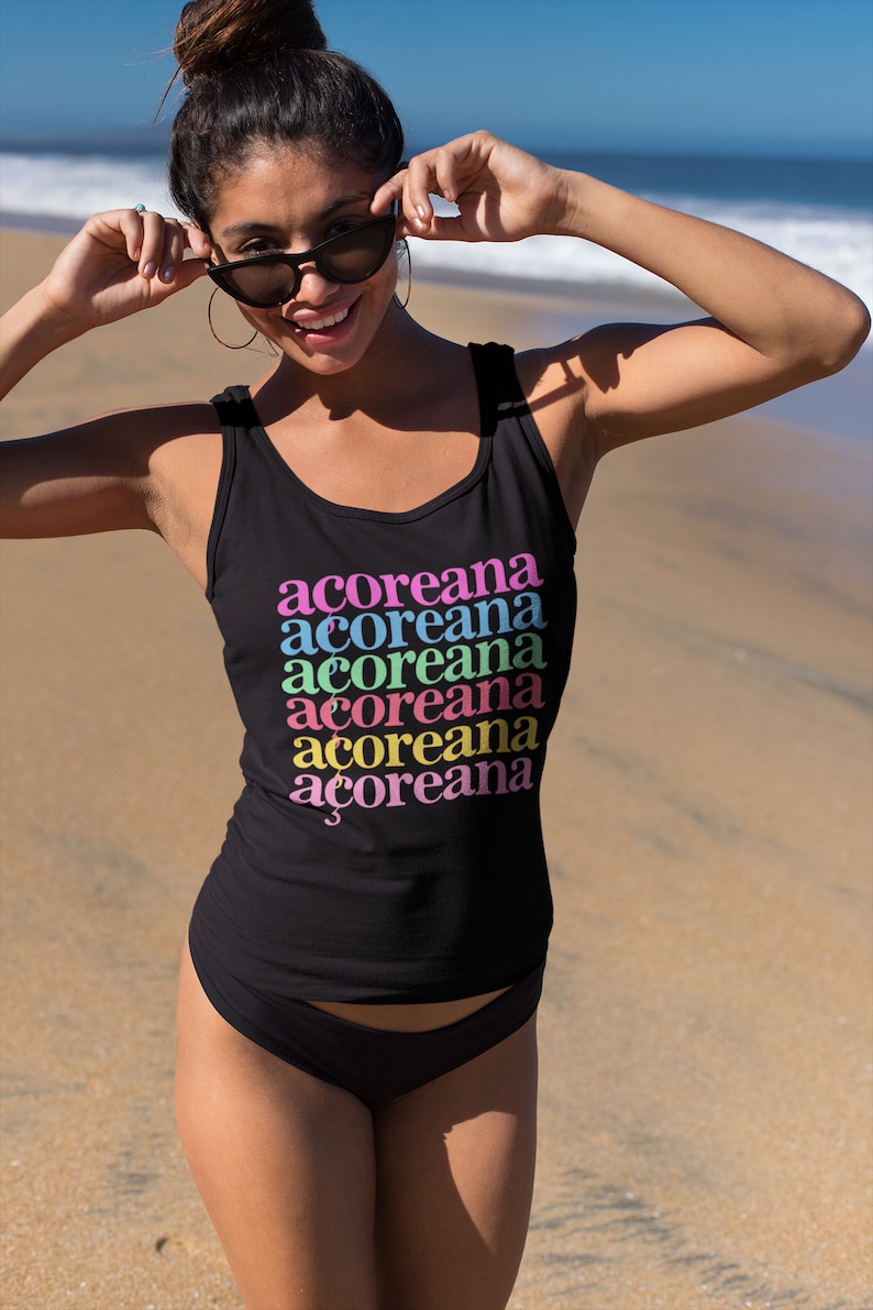 Op de afbeelding: Een zwart tanktop met het woord "acoreana" zes keer herhaald in verschillende kleuren: roze, blauw, groen, geel, oranje en roze. Het tanktop wordt gedragen door een persoon op een strand.