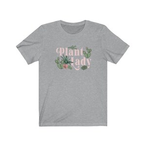 May include: A heather grey t-shirt with a pink graphic that says "Plant Lady" with a variety of green cacti and succulents surrounding the text.