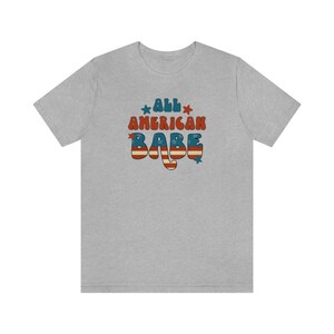 May include: A heather grey t-shirt with a retro-style graphic that says "All American Babe" in red, white, and blue with stars.