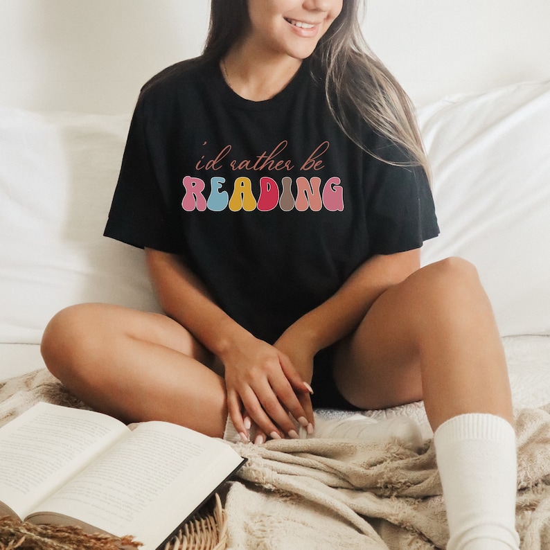 Puede incluir: Una camiseta negra con el texto "i'd rather be READING" en letras de colores de estilo retro. La camiseta la lleva puesta una persona sentada en una cama con un libro.