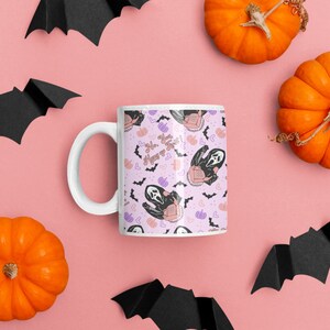 May include: White ceramic mug with a pink background and a repeating pattern of a ghost wearing a mask, bats, hearts, and pumpkins. The text "So, you wanna play?" is printed on the mug.