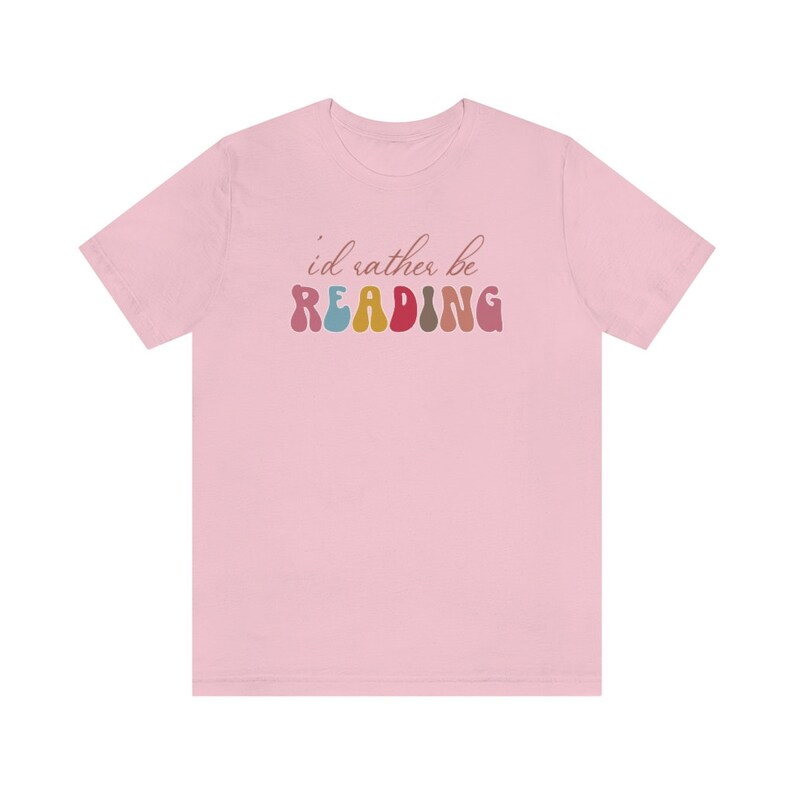 Puede incluir: Camiseta rosa con el texto "i'd rather be READING" en letras coloridas y onduladas.