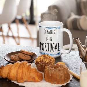 May include: White ceramic mug with a blue and white patterned design. The mug has the text "I'd rather be in Portugal" printed on it. The mug is sitting on a table with a plate of cookies, a croissant, and muffins.