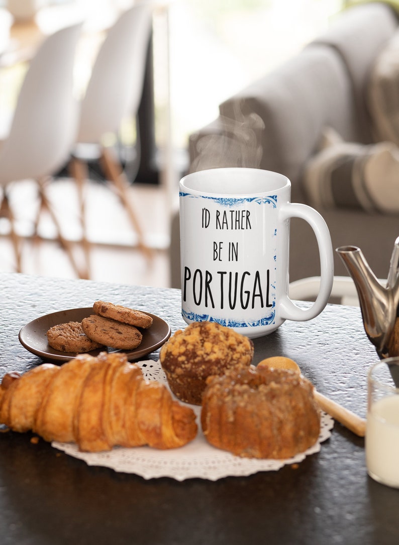 May include: White ceramic mug with a blue and white patterned design. The mug has the text "I'd rather be in Portugal" printed on it. The mug is sitting on a table with a plate of cookies, a croissant, and muffins.