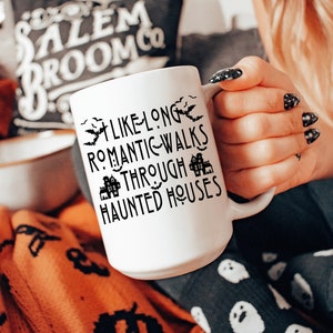 May include: White ceramic mug with black text that reads "Like long romantic walks through haunted houses". The mug is decorated with black bats and houses. The mug is being held by a hand wearing black nail polish.