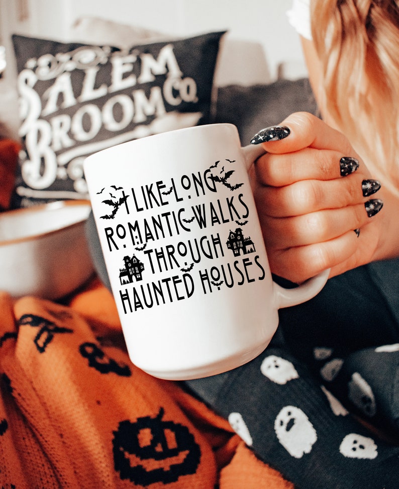 May include: White ceramic mug with black text that reads "Like long romantic walks through haunted houses". The mug is decorated with black bats and houses. The mug is being held by a hand wearing black nail polish.
