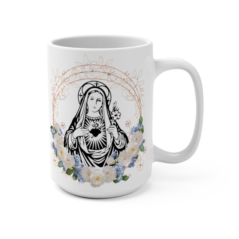 May include: White ceramic mug with a black and white image of the Virgin Mary with a heart in her chest. The image is surrounded by a wreath of white roses and blue flowers with gold accents.