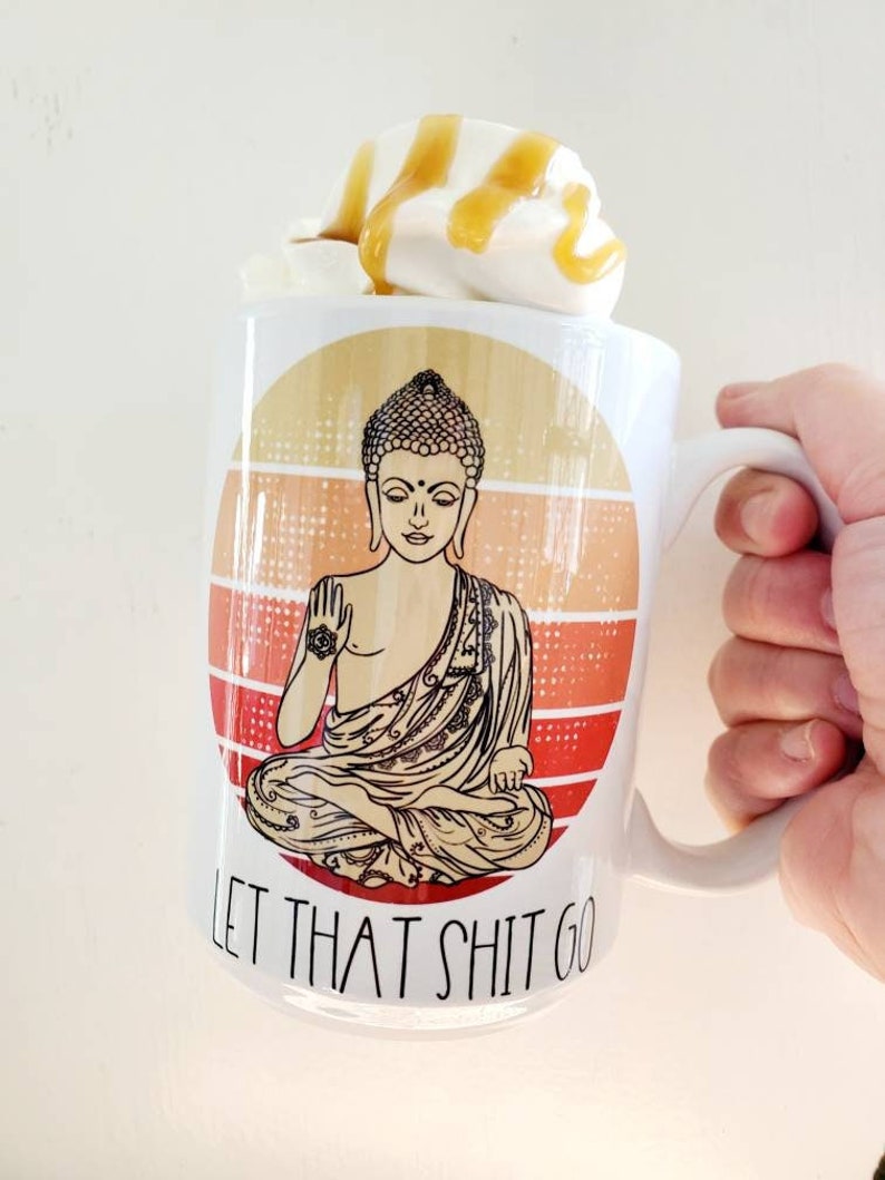 K&ouml;nnte beinhalten: Eine wei&szlig;e Keramiktasse mit einem Retro-Bild eines meditierenden Buddha mit dem Text "Let That Shit Go". Die Tasse ist gef&uuml;llt mit einem wei&szlig;en Getr&auml;nk, das mit Schlagsahne und Karamell betr&auml;ufelt ist.