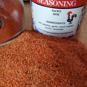 May include: A glass jar filled with red and orange Portuguese seasoning. The label on the jar reads "Portuguese Seasoning" and lists the ingredients: salt, paprika, garlic, black pepper, cumin, and coriander. The net weight is 14 oz.