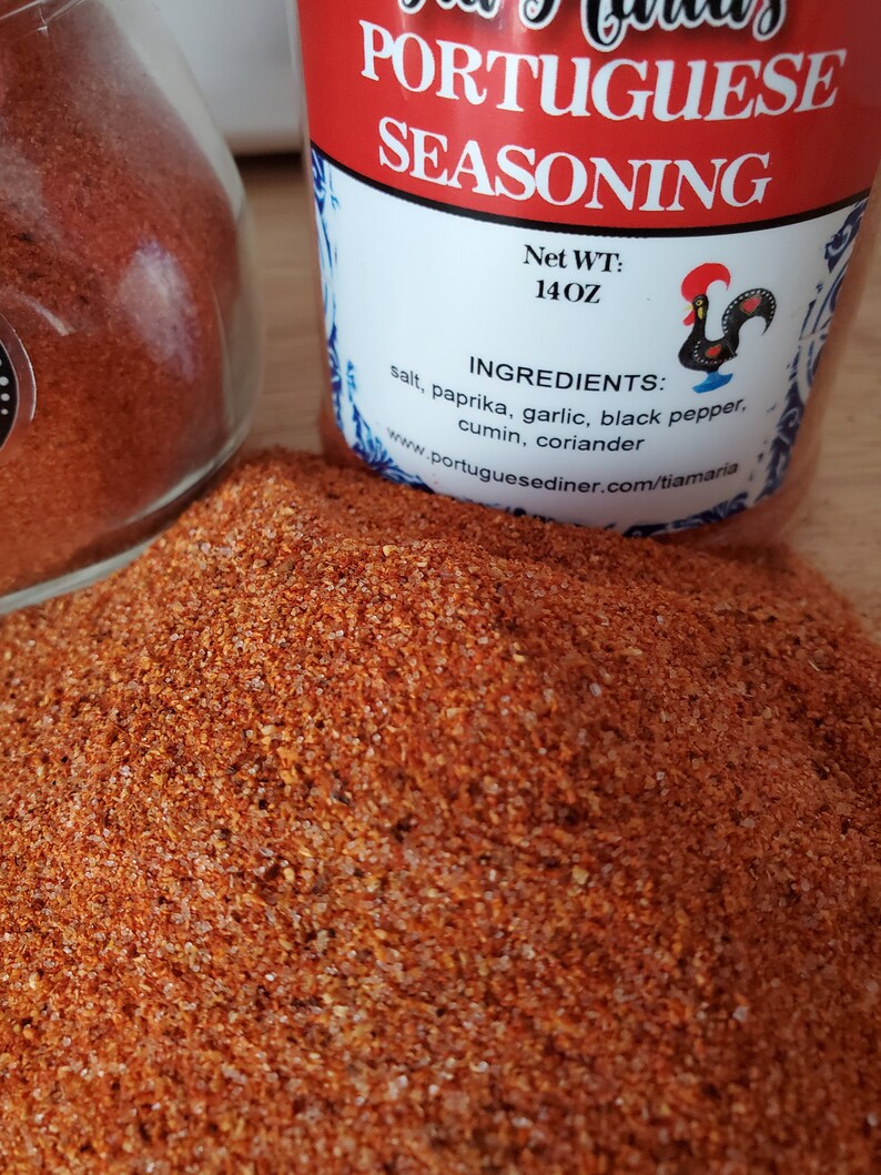May include: A glass jar filled with red and orange Portuguese seasoning. The label on the jar reads "Portuguese Seasoning" and lists the ingredients: salt, paprika, garlic, black pepper, cumin, and coriander. The net weight is 14 oz.