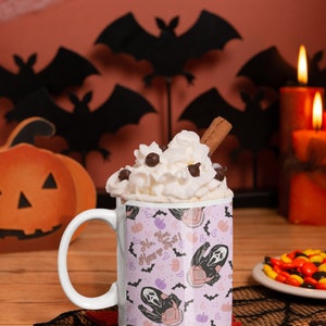 May include: White ceramic mug with a pink and purple pattern featuring a ghost holding a phone and the text "No. You. Text! Hang up". The mug is filled with whipped cream and a cinnamon stick. The mug is sitting on a black lace doily with a plate of candy corn in the background.