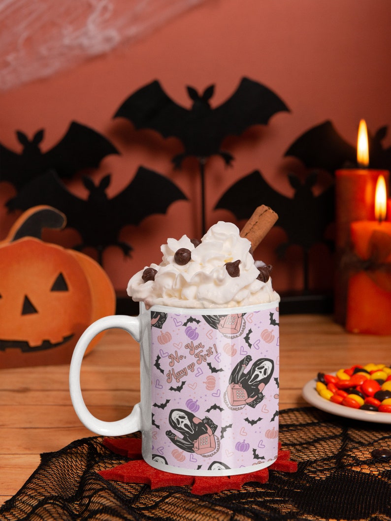 May include: White ceramic mug with a pink and purple pattern featuring a ghost holding a phone and the text "No. You. Text! Hang up". The mug is filled with whipped cream and a cinnamon stick. The mug is sitting on a black lace doily with a plate of candy corn in the background.
