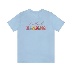 Puede incluir: Camiseta azul claro con el texto "i'd rather be READING" en letras de colores de estilo retro.