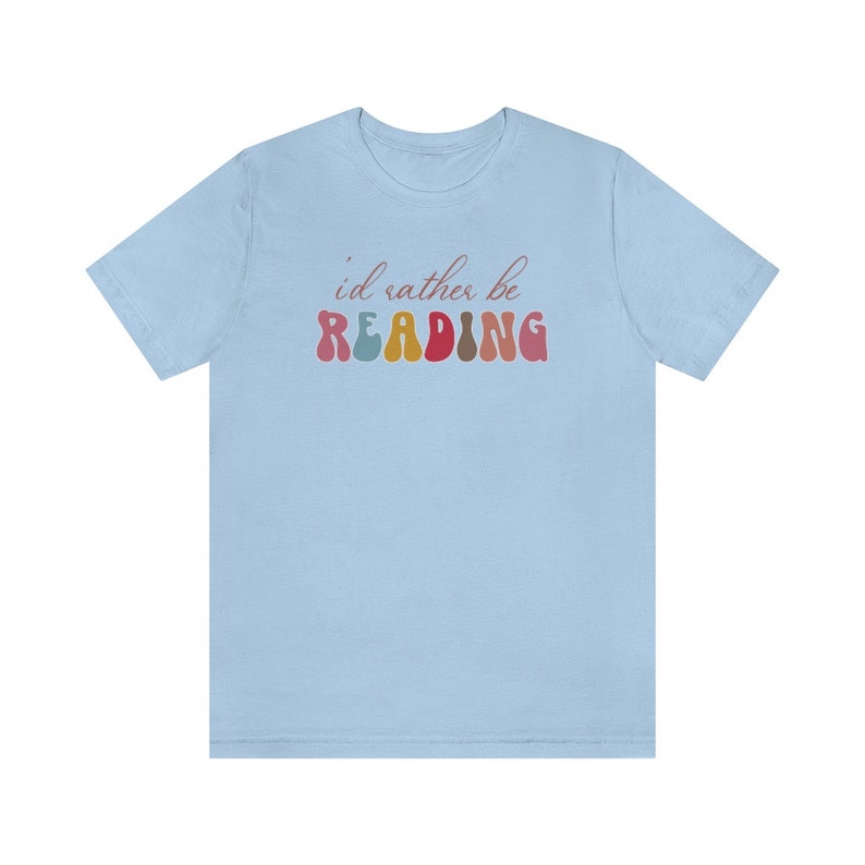 Puede incluir: Camiseta azul claro con el texto "i'd rather be READING" en letras de colores de estilo retro.