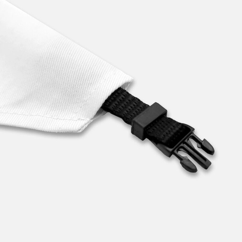 May include: Close-up of a white fabric corner with a black nylon strap and a black plastic buckle. The buckle has three prongs for secure fastening. The fabric appears to be a sturdy, woven material.
