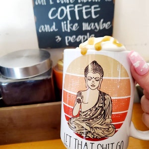 K&ouml;nnte beinhalten: Eine wei&szlig;e Keramik-Kaffeetasse mit einem Grafikdruck eines meditierenden Buddha vor einem Sonnenuntergangs-Hintergrund. Die Tasse hat den Text "LET THAT SHIT GO" auf dem Boden aufgedruckt. Die Tasse ist mit Kaffee gef&uuml;llt und mit Schlagsahne und Karamellso&szlig;e garniert.