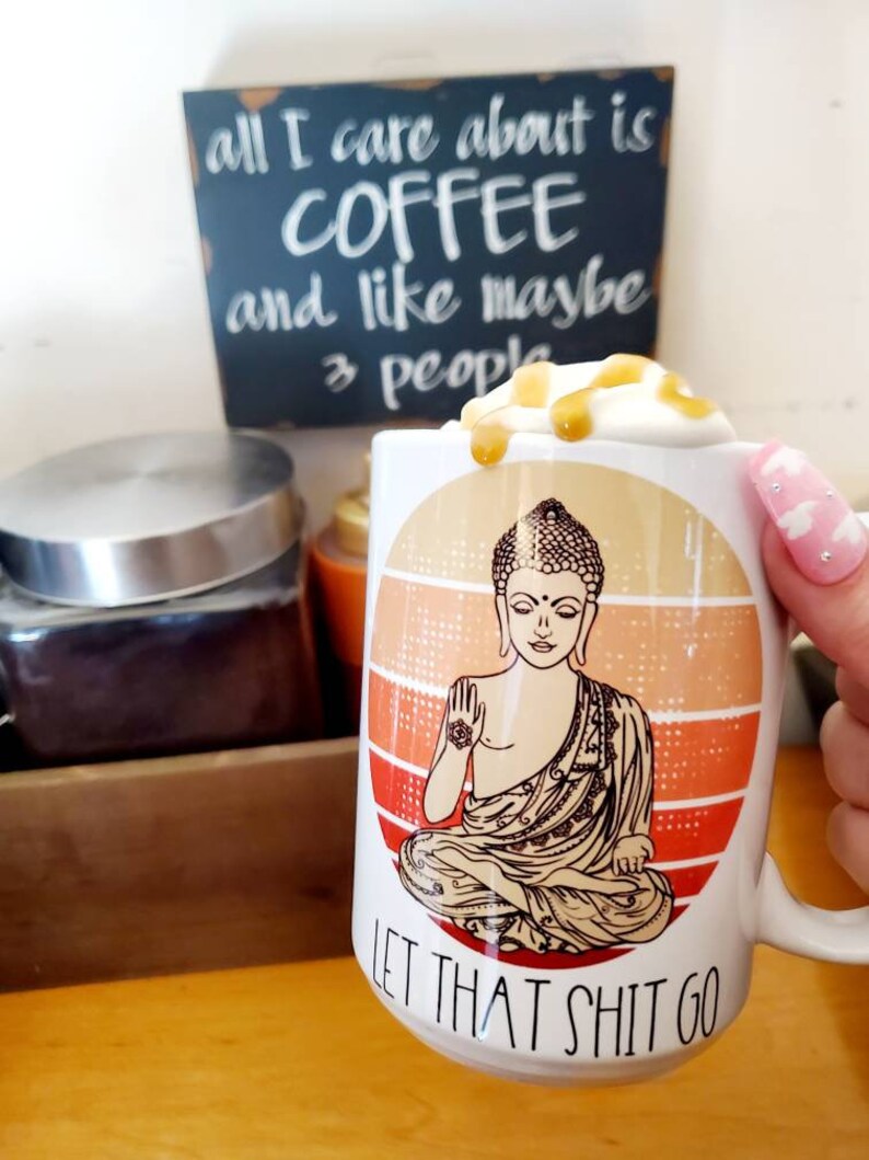 K&ouml;nnte beinhalten: Eine wei&szlig;e Keramik-Kaffeetasse mit einem Grafikdruck eines meditierenden Buddha vor einem Sonnenuntergangs-Hintergrund. Die Tasse hat den Text "LET THAT SHIT GO" auf dem Boden aufgedruckt. Die Tasse ist mit Kaffee gef&uuml;llt und mit Schlagsahne und Karamellso&szlig;e garniert.