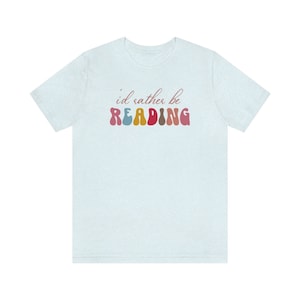 Puede incluir: Una camiseta azul claro con el texto "i'd rather be READING" en letras de colores del arco&iacute;ris.