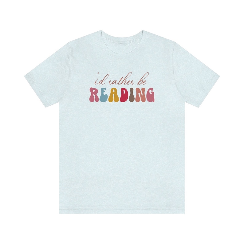 Puede incluir: Una camiseta azul claro con el texto "i'd rather be READING" en letras de colores del arco&iacute;ris.