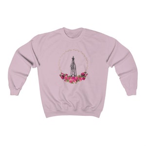 Pu&ograve; includere: Una felpa rosa con una grafica di Nostra Signora di Fatima circondata da una ghirlanda di rose rosa.