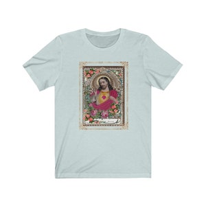 May include: A light blue t-shirt with a vintage image of Jesus Christ in a gold frame with floral accents. The image is surrounded by pink roses and the word "Jesus" is printed below the image.