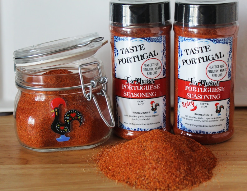 May include: Two glass jars of Tia Maria's Portuguese Seasoning, one labeled "Spicy". The jars are filled with a red-orange powder and have a rooster logo on the front. The jars are sitting on a wooden surface with a pile of the seasoning in front of them.