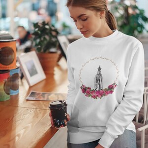 Pu&ograve; includere: Felpa bianca con una grafica di Nostra Signora di Fatima in una ghirlanda di rose rosa. La grafica &egrave; in bianco e nero.