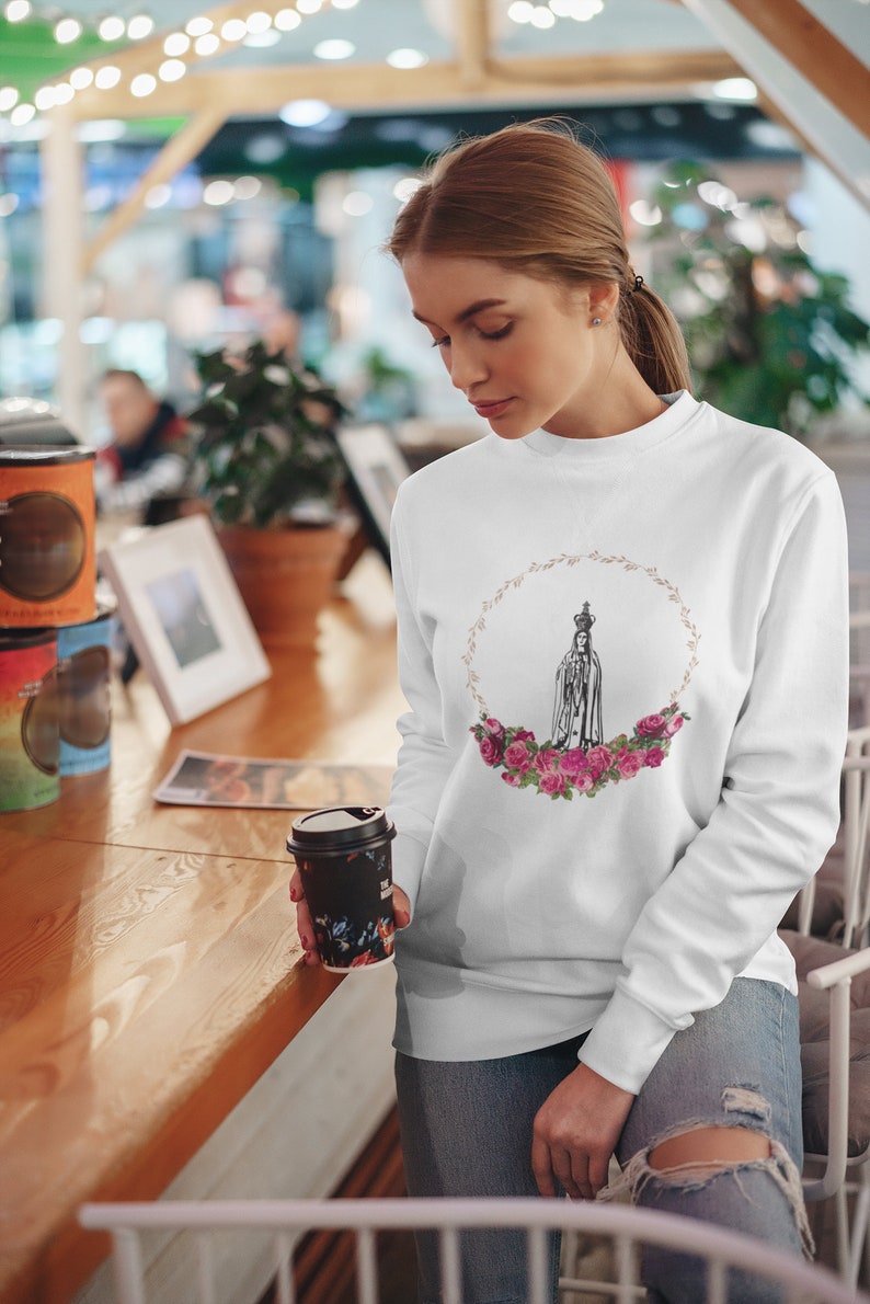 Pu&ograve; includere: Felpa bianca con una grafica di Nostra Signora di Fatima in una ghirlanda di rose rosa. La grafica &egrave; in bianco e nero.