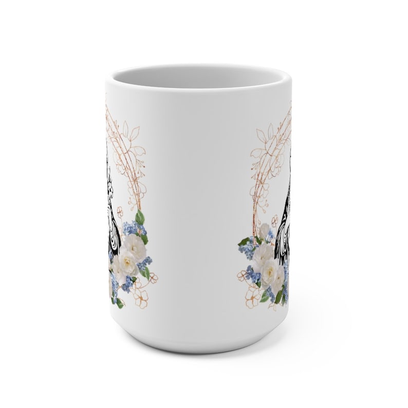 May include: White ceramic mug with a black and white floral design. The design features a floral wreath with white roses and blue flowers. The wreath is outlined in a gold color.