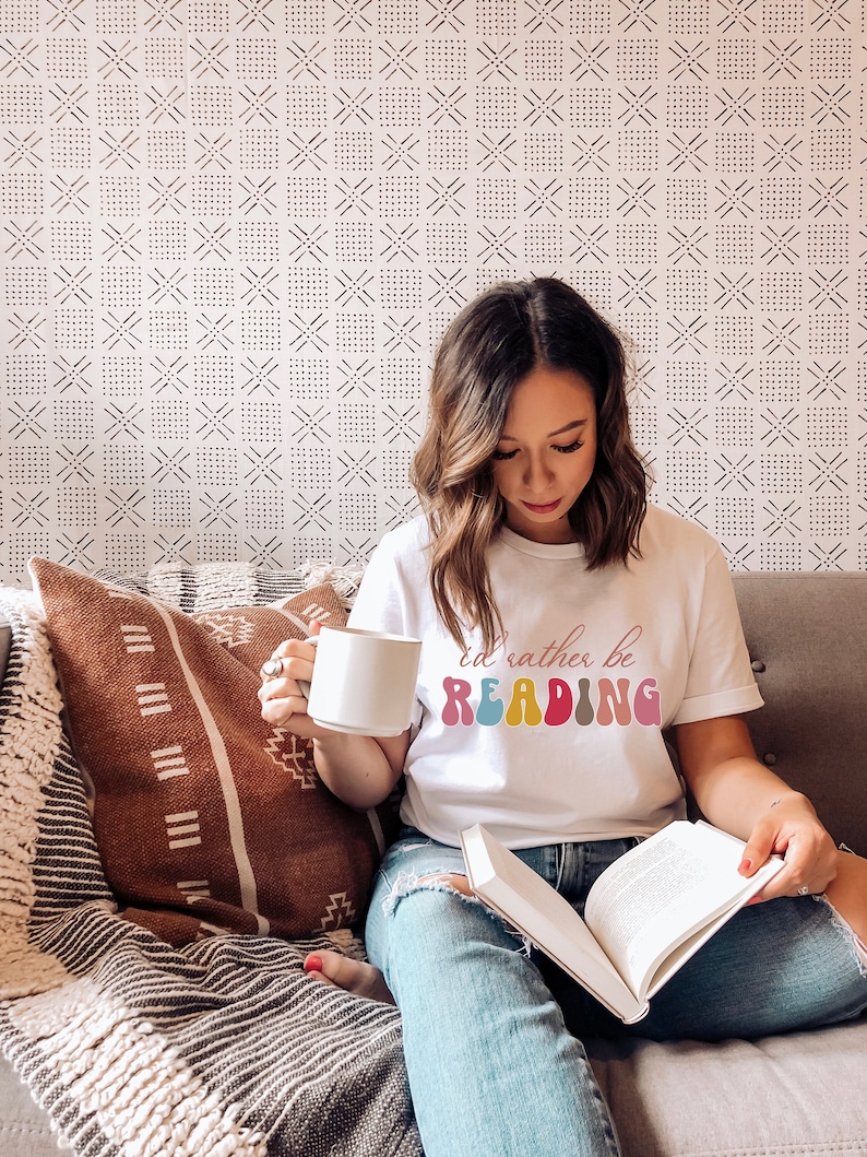 Puede incluir: Una mujer con una camiseta blanca con el texto "I'd rather be READING" en letras de colores, sentada en un sof&aacute; con un libro en su regazo y una taza blanca en su mano.