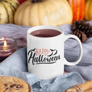 Może przedstawiać: Biały ceramiczny kubek z czarnym i pomarańczowym nadrukiem "Happy Halloween" i nietoperzami. Kubek jest wypełniony ciemnobrązowym płynem i stoi na niebieskim dzianym kocu z cynamonowymi patyczkami i szyszką.