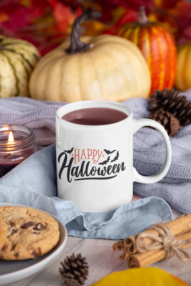 Może przedstawiać: Biały ceramiczny kubek z czarnym i pomarańczowym nadrukiem "Happy Halloween" i nietoperzami. Kubek jest wypełniony ciemnobrązowym płynem i stoi na niebieskim dzianym kocu z cynamonowymi patyczkami i szyszką.