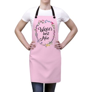 May include: Pink apron with adjustable straps and a floral wreath design. The wreath has the text "World's best M&atilde;e" in black.