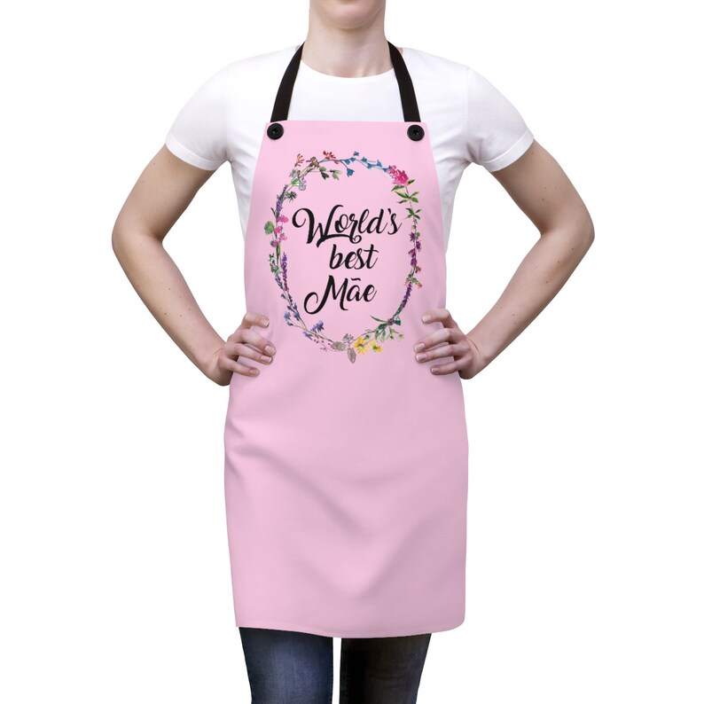 May include: Pink apron with adjustable straps and a floral wreath design. The wreath has the text "World's best M&atilde;e" in black.