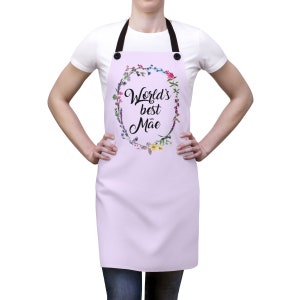 May include: A lavender apron with adjustable straps and a floral wreath design. The wreath features colorful flowers and the text "World's best M&atilde;e".