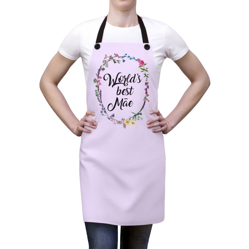 May include: A lavender apron with adjustable straps and a floral wreath design. The wreath features colorful flowers and the text "World's best M&atilde;e".
