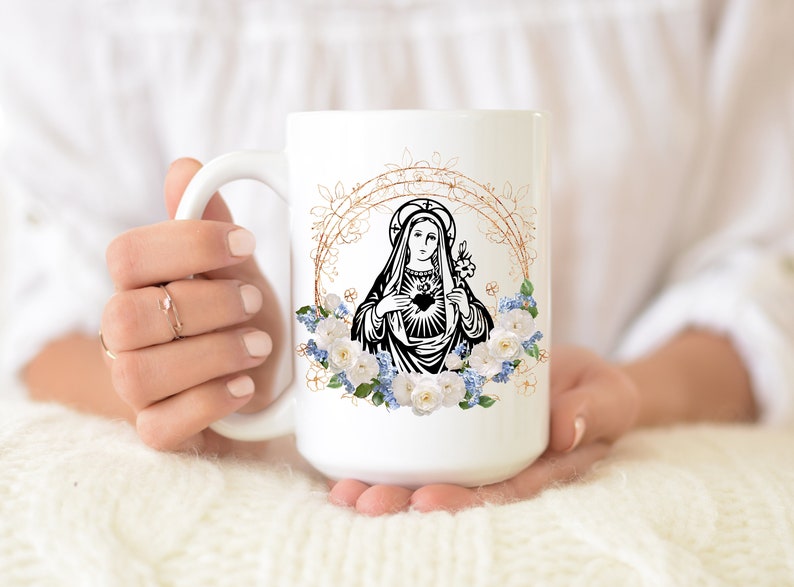 May include: White ceramic mug with a black and white image of the Virgin Mary with a heart in her chest. The image is surrounded by a gold floral wreath with white and blue flowers.