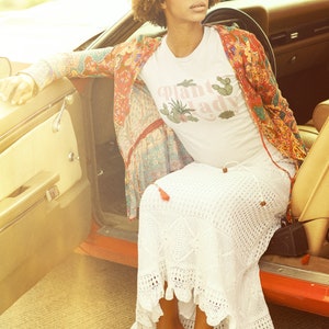 May include: A woman wearing a white t-shirt with the text "Plant Lady" in pink lettering, a red and floral patterned jacket, a white crochet skirt, and brown sandals. She is sitting in a vintage car with the door open.