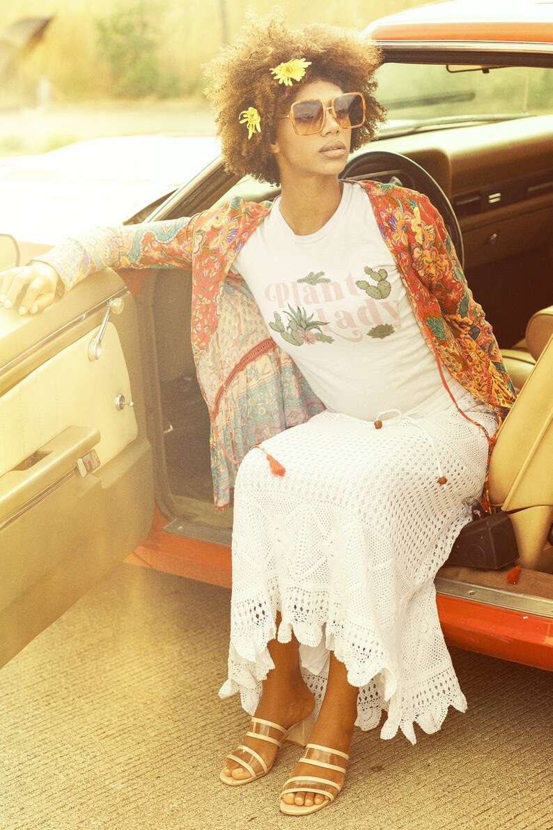 May include: A woman wearing a white t-shirt with the text "Plant Lady" in pink lettering, a red and floral patterned jacket, a white crochet skirt, and brown sandals. She is sitting in a vintage car with the door open.