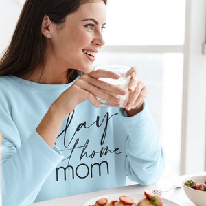 May include: A woman wearing a light blue sweatshirt with the text "Stay Home Mom" in black lettering. She is holding a glass of milk and smiling.